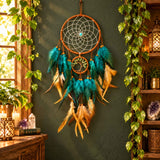 Teal and gold feather dreamcatcher with a hanging Tree of Life hoop on a dark green wall, framed by pothos vines, lanterns and crystals.
